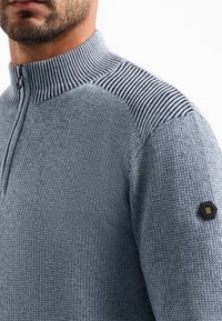 Close-up van een man in een lichtblauwe gebreide trui met een kwartrits, geribde kraag, gestreept schouderpatroon en hexagonaal mouwpatch.