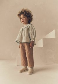 Vit blus med rutigt mönster och gröna detaljer, beige kordfärgade byxor och beiga skor. Barnet står framför en ljus bakgrund med trappor.