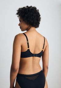 Black bra with adjustable straps, gold hardware accents, and a hook-and-eye closure. Features a smooth texture and minimalistic design.