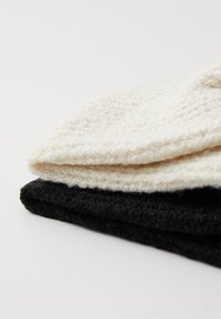 Due cappelli di lana impilati; uno bianco e uno nero. Il cappello bianco ha un motivo testurizzato, mentre il cappello nero è liscio e denso.