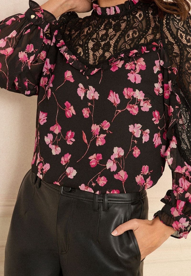 Femme portant un chemisier noir à fleurs roses avec un yoke en dentelle, associé à un pantalon en cuir noir, main dans la poche.