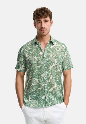 Młody mężczyzna o kręconych włosach nosi zieloną i białą koszulę w tropikalne liście z krótkim rękawem oraz białe spodnie, ma ręce w kieszeniach, tło w neutralnym kolorze.