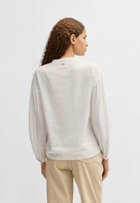 Blusa bianca con motivo a pois testurizzati, maniche lunghe, polsi arricciati e scollatura rotonda. La modella mostra la vista posteriore.