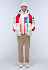 Giubbotto imbottito in crema, rosso e blu con cappuccio, indossato sopra un top in pile bianco; pantaloni beige e sneakers bianche completano il look.
