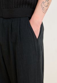 Black linen pants feature a relaxed fit with side pockets, soft texture, and subtle vertical pleats, complemented by a ribbed black knit sweater.