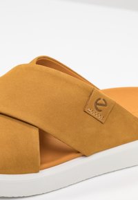 Sandale à enfiler en suède beige avec des straps croisées, une semelle intérieure douce de teinte plus claire et une étiquette de logo discrète avec des détails de couture.