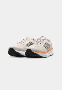 Beige sportsko med en mesh-overdel, orange mellemsål og en struktureret sort gummisål. Har et fremtrædende logo og snørebåndsdesign.