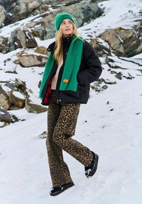 Žena v zelené čepici a šále, černé péřové bundě, kalhotách s leopardím potiskem a černých botách stojící na zasněženém skalnatém terénu.