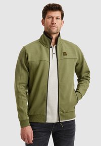 Olijfgroene jas met hoge kraag, gestructureerde schouderpanelen, ritsluiting en zakken aan de zijkant, gecombineerd met een lichtgrijs shirt eronder.