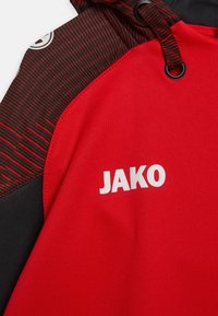 Rode hoodie met zwarte accenten, gestructureerd ontwerp en een opvallend wit "JAKO"-logo. Materialen omvatten een stof met een gladde textuur.