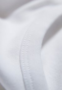 Nahaufnahme von weißem Stoff, der eine glatte Textur und eine geschwungene Naht mit feinen Stichdetails zeigt. Das Material wirkt weich und leicht.
