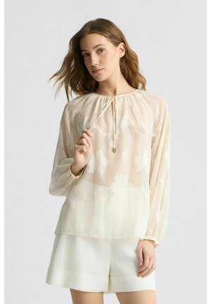 Blusa cremosa trasparente con motivo floreale, scollatura arricciata, maniche lunghe e una texture sottile. Abbinata a pantaloni corti bianchi.