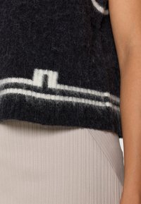 Top preto de lã com padrão geométrico branco, textura felpuda. Usado sobre um tecido beige claro texturizado com pregas e uma costura em ângulo.