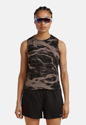 Person mit geflochtenem Haar trägt ein ärmelloses Oberteil mit schwarzem und braunem Muster, schwarze Shorts und reflektierende Sportsonnenbrille, schaut nach vorne.