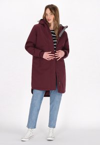 Cappotto impermeabile color bordeaux con cappuccio, chiusura con zip e due tasche laterali. Indossato sopra un top a righe bianche e nere e jeans blu chiaro.