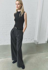 Haut sans manches noir associé à un pantalon gris à motifs plissés taille haute. Comprend une ceinture noire et des chaussures noires pointues. Fond neutre.