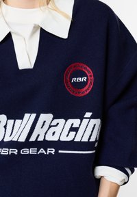 Άτομο που φοράει μπλε σκούνο πουλόβερ Red Bull Racing με λευκό γιακαδάκι και μανσέτες μανικιών, με λογότυπα και κείμενα Red Bull Racing RBR στο στήθος.