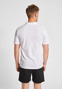 White short-sleeve polo shirt with a solid texture, featuring a collar and a straight hem, worn with black shorts. View from the back.