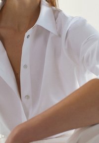 Chemise blanche avec un col classique, dotée d’une patte de boutonnage et d’un tissu lisse. Bras reposant sur une surface douce, mettant en valeur des lignes épurées.