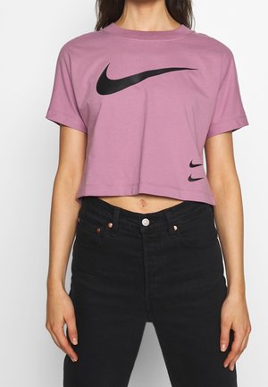 Žena nosící krátké fialové tričko Nike s velkými a malými černými logy Nike, doplněné o černé džíny s vysokým pasem.