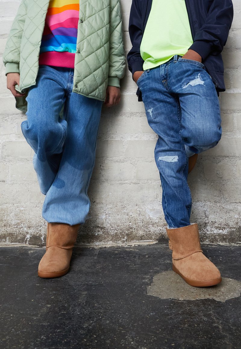 Deux paires de jambes contre un mur, portant des jeans bleus usés et des bottes en daim beige. Une personne porte une veste matelassée verte et un pull rayé multicolore.