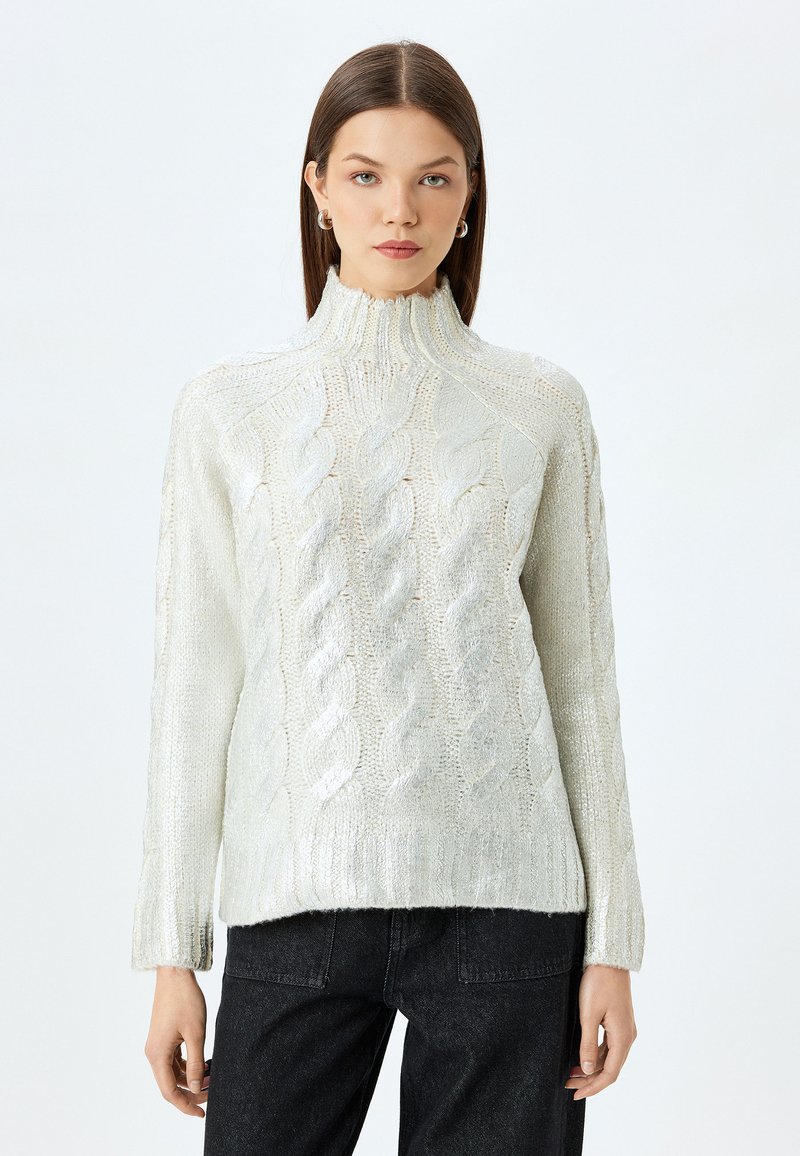 Hellgrauer Kabelstrickpullover mit hohem Kragen und gerippten Bündchen, der ein strukturiertes Muster und eine lockere Passform aufweist, kombiniert mit dunklen Jeans.