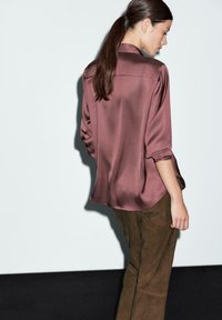 Camicia di satin in malva con una vestibilità rilassata, dotata di maniche arrotolate e un yoke posteriore, abbinata a pantaloni in suede marroni.