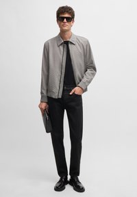 Gray patterned zip-up jacket, black t-shirt, black jeans, and black shoes. Holding a black clutch. Curly hair and sunglasses.