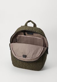 Sac à dos en tissu matelassé vert avec une ouverture zippée. Intérieur beige avec un compartiment divisé. Dispose d'une poignée supérieure pour le transport.