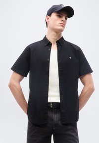 Νέος άνδρας που φοράει μαύρο πουκάμισο με κοντά μανίκια και κουμπιά πάνω από λευκό αμάνικο, μαύρο τζιν, μαύρη ζώνη και μαύρο καπέλο μπέιζμπολ, με τα χέρια πίσω από την πλάτη.