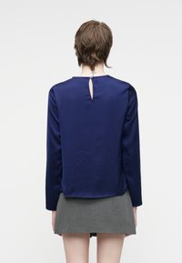 Persona con capelli corti castani che indossa una blusa a maniche lunghe blu navy con un bottone a chiave sulla schiena e una mini gonna grigia, rivolta di spalle.