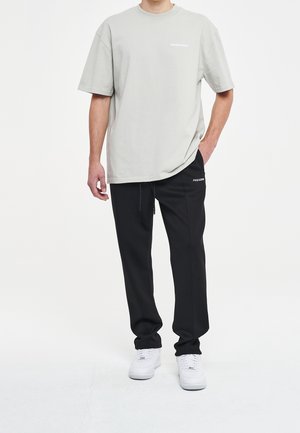 Helles graues Baumwoll-T-Shirt kombiniert mit schwarzen Jogginghosen. Das T-Shirt hat ein dezentes Logo; die Jogginghosen verfügen über einen Kordelzug und Logo-Details. Weiße Sneaker.