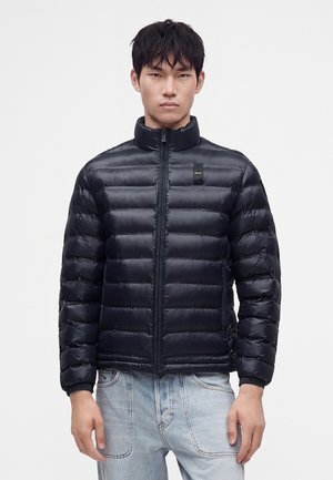 Mann trägt eine schwarze gesteppte Daunenjacke mit Frontreißverschluss und hellblauen Jeans, steht vor einem schlichten weißen Hintergrund.