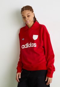 Felpa rossa in cotone con colletto, logo Adidas bianco e iconiche tre strisce azzurre. Pantaloni neri con tasche.