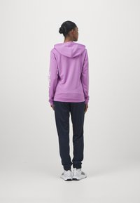 Fialová mikina s kapucňou a bielym logom na rukáve, skombinovaná s námorníckymi jogger nohavicami a bielymi športovými topánkami, pohľad zozadu.