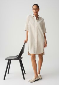 Beige Hemdkleid mit kurzen Ärmeln, Knopfleiste und zwei Brusttaschen. Gestylt mit beigen Slippern und neben einem schwarzen Stuhl platziert.