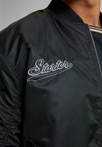Veste bomber noire avec le logo blanc "Starter" brodé sur le côté gauche de la poitrine, portée sur une chemise à col rayé.