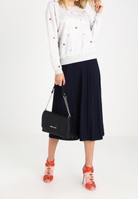 Donna che indossa un maglione grigio con pois colorati, una gonna midi plissettata blu navy, scarpe con tacco rosso aperte e porta una borsa nera con tracolla a catena.