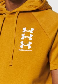 Felpa gialla con cappuccio, caratterizzata da tre loghi bianchi Under Armour sul davanti. Tessuto liscio e maniche corte.