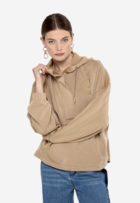 Sudadera beige de tela suave, con bolsillo frontal, cordón ajustable y detalle de logo en relieve. Combinada con jeans azules.