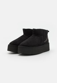 Une paire de bottines en daim noires avec semelles épaisses et couture visible à l'arrière, présentées sur un fond blanc.