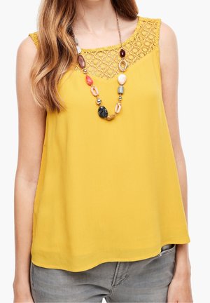 Vrouw draagt een mouwloze gele top met kanten halslijn en een meerkleurige kralenketting, gecombineerd met een grijze broek.