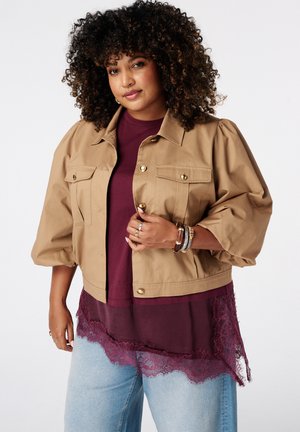 Femme aux cheveux bouclés portant une veste beige sur un top bordeaux orné de dentelle et un jean bleu clair, accessoirisée de bracelets et de bagues.