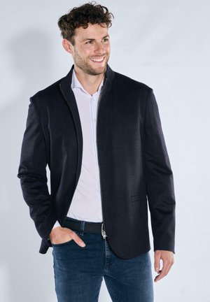 Lächelnder Mann, der ein schwarzes, zum Reißverschluss geöffnetes Sakko, ein weißes Hemd und dunkle Jeans trägt, mit einer Hand in der Tasche, stehend vor einem einfarbigen Hintergrund.