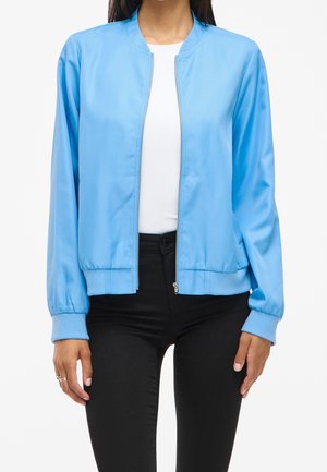Femme portant une veste bomber bleu clair zippée par-dessus une chemise blanche et un pantalon noir, debout devant un fond uni.
