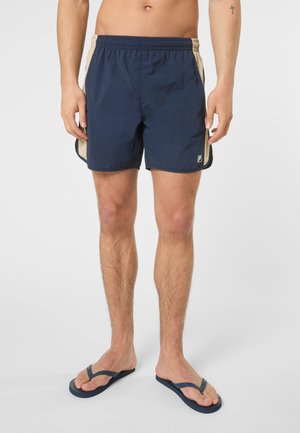 Uomo che indossa pantaloncini sportivi blu navy con pannelli laterali beige e infradito blu navy, in piedi contro uno sfondo grigio uniforme.