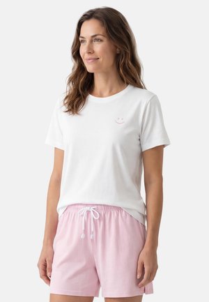 Kvinde med bølget brunt hår iført en hvid t-shirt med et lille lyserødt smil og lyserøde shorts med snøre på hvid baggrund.
