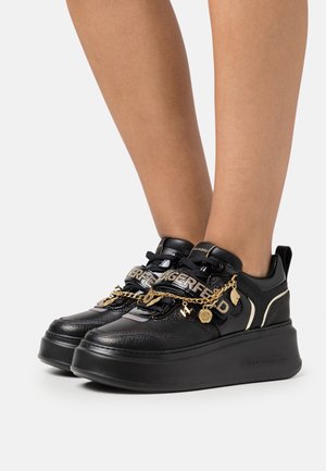 Baskets plateforme noires avec chaîne dorée et breloques en lettres, avec le texte « LAGERFELD » sur les sangles, portées sur des jambes nues sur un fond uni.