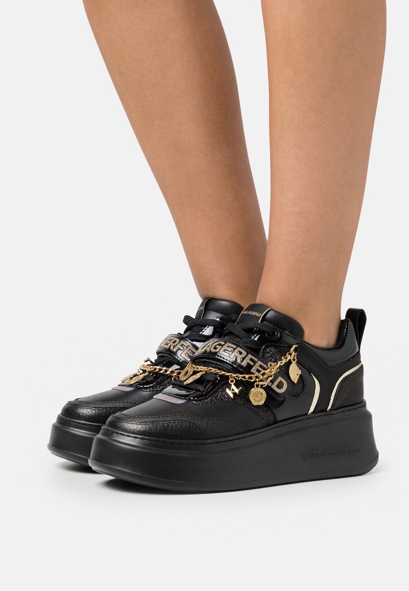 Baskets plateforme noires avec chaîne dorée et breloques en lettres, avec le texte « LAGERFELD » sur les sangles, portées sur des jambes nues sur un fond uni.