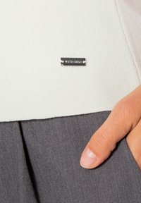 Bílý pletený top s šedým skládáním ve spodní části a stříbrnou štítkovou logem. Detailní záběr ruky, která ukazuje na štítek na oděvu.